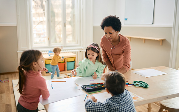 Kinder spielen und malen an einem Tisch neben einer Erzieherin.
